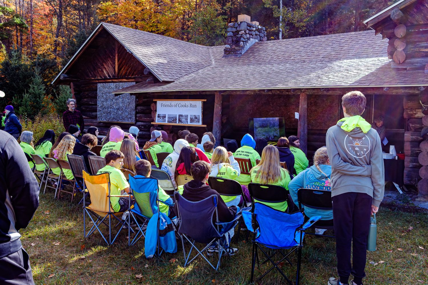 Students in green event shirts seated on chairs in front of the cabin, watching a film on a screen on the porch