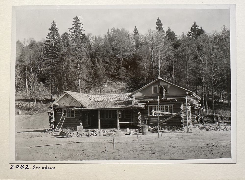 The log cabin under construction with a crewman on the roof ridge