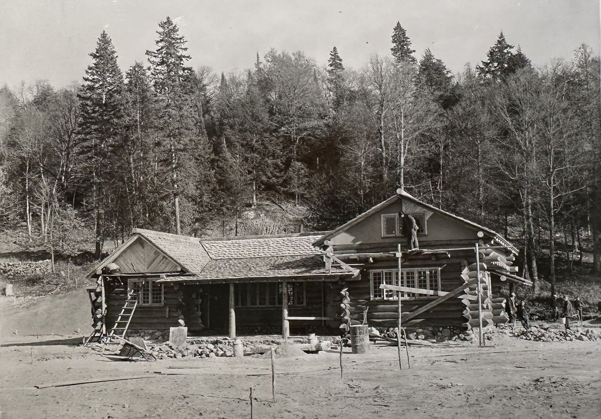 Black-and-white 1935 photograph of the log cabin under construction, a crewman on the roof ridge